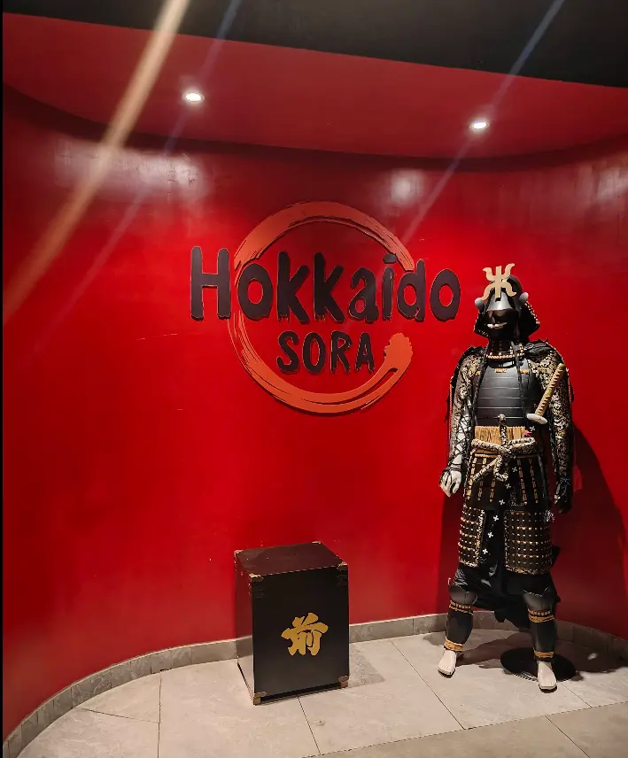 A suit of Japanese samurai armour on display against the deep vermilion wall of Hokkaido Sora.